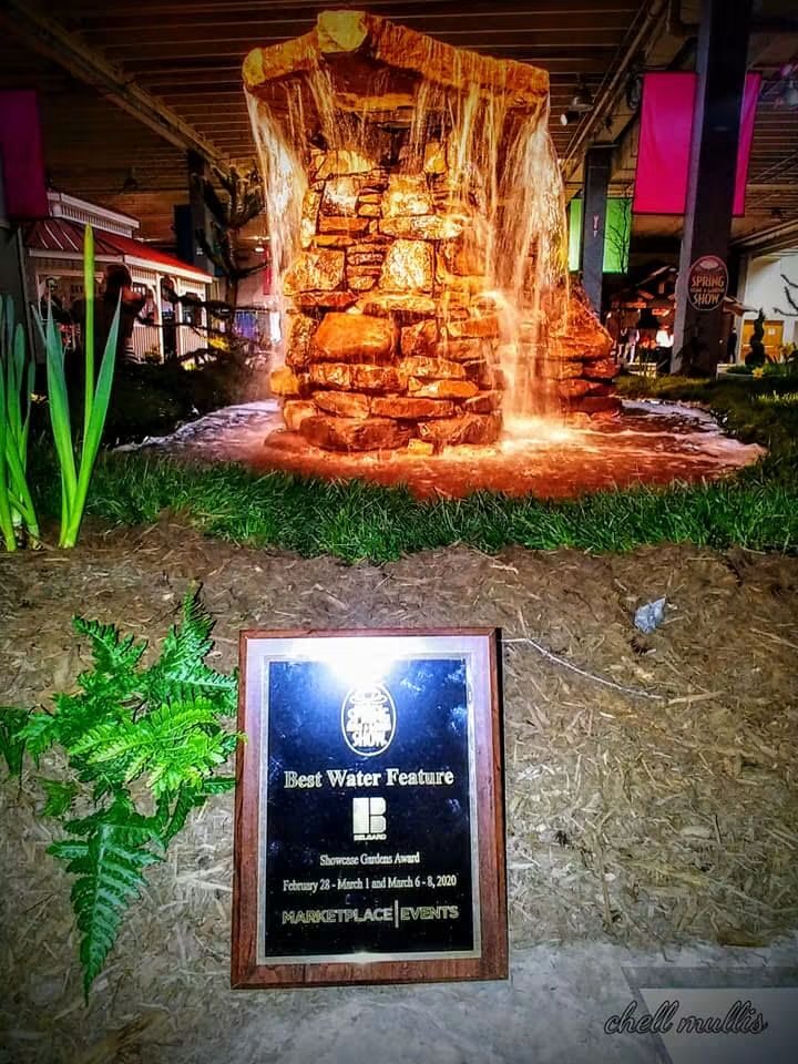 A Best Water Feature award plaque in front of a lit stacked-stone waterfall at a garden show
