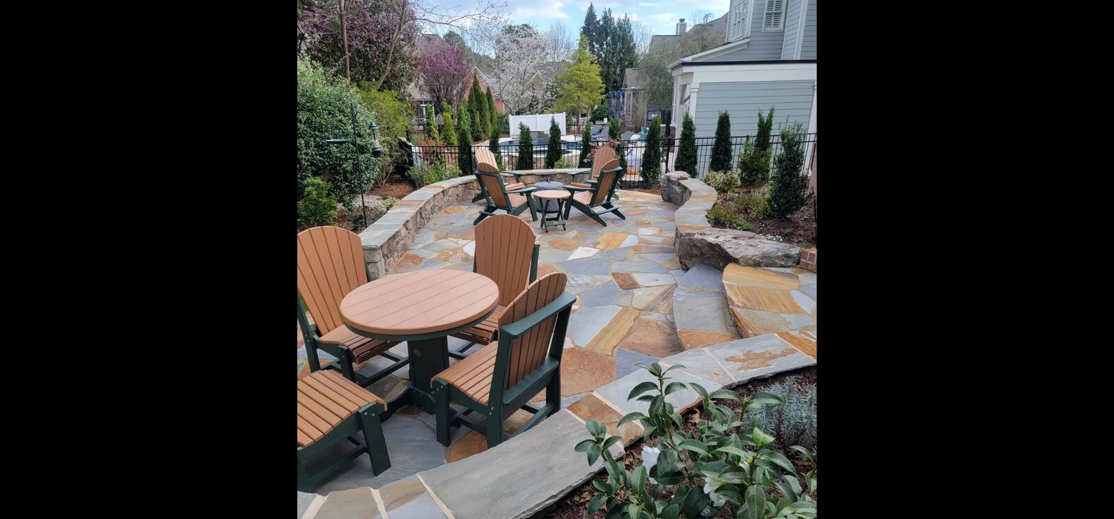 A flagstone patio with Adirondack chairs around a fire pit and dining table, overlooking a pool beyond a stacked-stone wall