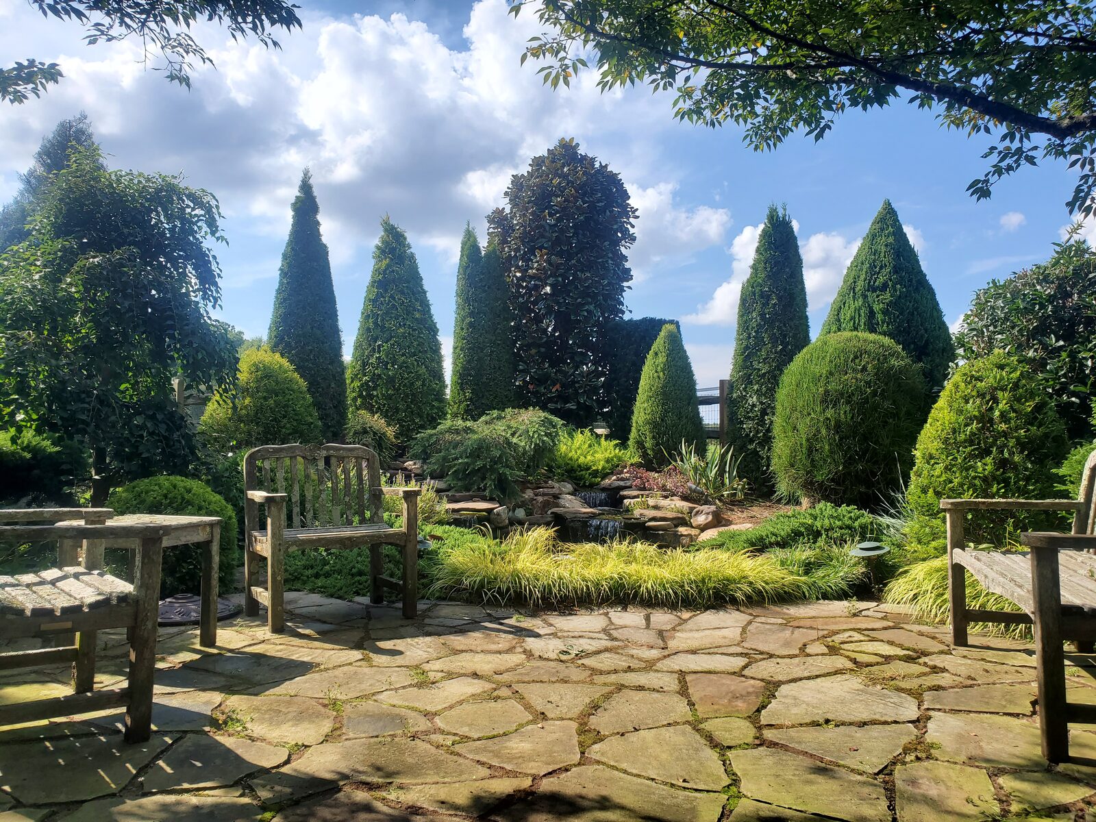 A shaded flagstone patio with wooden garden benches beside a small stream and pond, framed by mature conifers