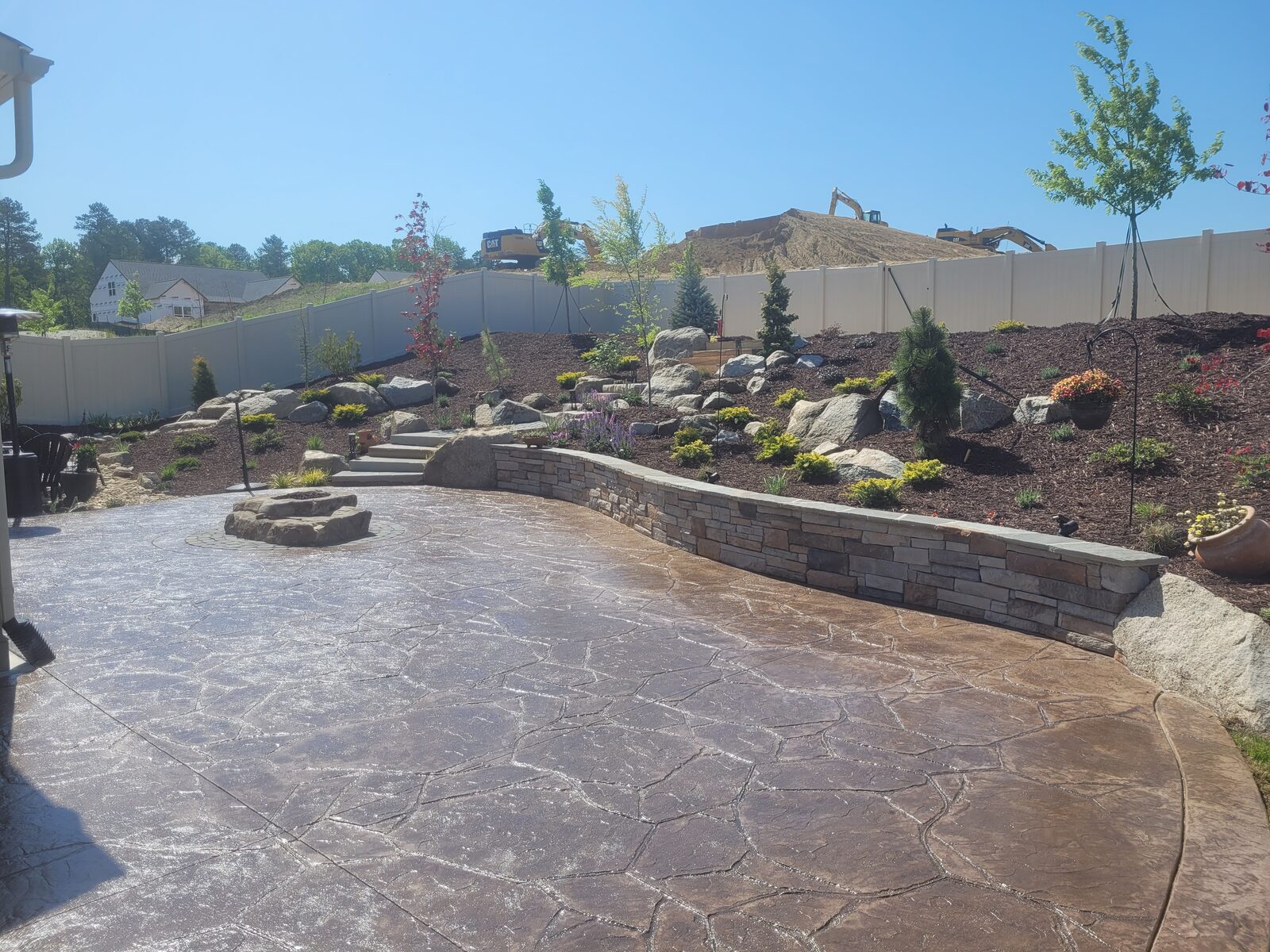 A curved stamped-concrete patio with a fire pit and stacked-stone seat wall, with grading equipment reshaping the hillside beyond